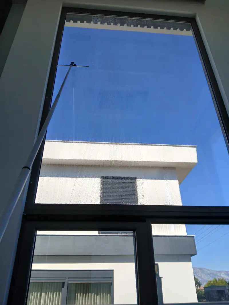Büyük pencere dış yüzeyinde sırıkla cam temizliği, mavi gökyüzü yansıması